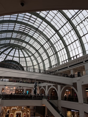 An interior of a shopping mall with a grand, arched glass ceiling that allows natural light to fill the space. The architecture includes ornate railings and multiple levels with escalators connecting the floors. Several luxury retail stores can be seen on the lower level.