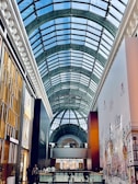 A modern indoor shopping mall with a large, arched glass ceiling allowing natural light to illuminate the space. Storefronts line the walkway, with a focus on Polo by Ralph Lauren. People are casually walking and socializing throughout the area.