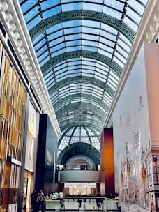 A modern indoor shopping mall with a large, arched glass ceiling allowing natural light to illuminate the space. Storefronts line the walkway, with a focus on Polo by Ralph Lauren. People are casually walking and socializing throughout the area.