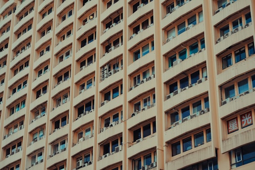 A tall building with multiple rows of windows, each equipped with air conditioning units. The facade is a beige color, and there are signs with Asian script on them, adding cultural context to the scene.