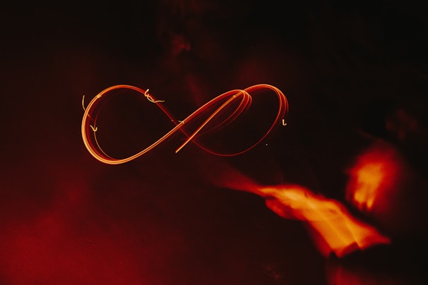 A glowing, fiery orange infinity symbol traced against a dark background, creating a sense of movement and fluidity. The light trails are vivid and contrast with the surrounding darkness, adding a dramatic effect.