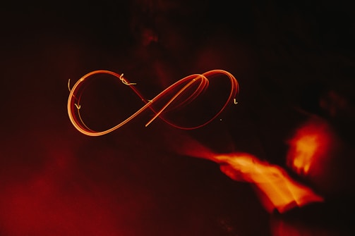 A glowing, fiery orange infinity symbol traced against a dark background, creating a sense of movement and fluidity. The light trails are vivid and contrast with the surrounding darkness, adding a dramatic effect.