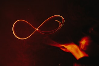 A glowing, red infinity symbol is drawn in mid-air against a dark background. It appears to be created by a light painting technique, with trails of light forming the continuous loop. On the right, there is a blurred trace of a hand motion, suggesting the movement that created the shape.