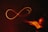 A glowing, red infinity symbol is drawn in mid-air against a dark background. It appears to be created by a light painting technique, with trails of light forming the continuous loop. On the right, there is a blurred trace of a hand motion, suggesting the movement that created the shape.