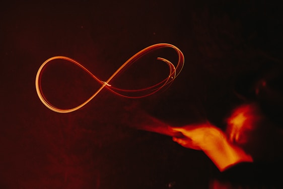 A glowing, red infinity symbol is drawn in mid-air against a dark background. It appears to be created by a light painting technique, with trails of light forming the continuous loop. On the right, there is a blurred trace of a hand motion, suggesting the movement that created the shape.