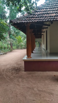 A traditional house with a tiled roof and wooden pillars, surrounded by lush green trees. The ground is sandy, and a wide verandah extends along the side of the house.