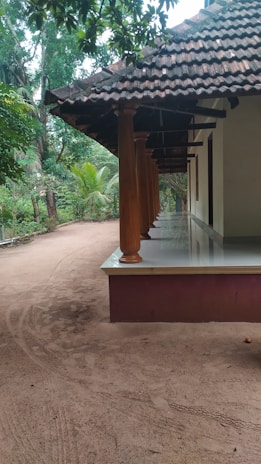 A traditional house with a tiled roof and wooden pillars, surrounded by lush green trees. The ground is sandy, and a wide verandah extends along the side of the house.