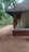 A traditional house with a tiled roof and wooden pillars, surrounded by lush green trees. The ground is sandy, and a wide verandah extends along the side of the house.