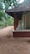 A traditional house with a tiled roof and wooden pillars, surrounded by lush green trees. The ground is sandy, and a wide verandah extends along the side of the house.