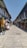 A narrow street lined with shops on both sides, selling a variety of clothing and souvenirs. The shops have colorful awnings and display racks with clothes hanging and other items. The street appears to be empty of people, with an expansive clear sky above.