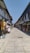 A narrow street lined with shops on both sides, selling a variety of clothing and souvenirs. The shops have colorful awnings and display racks with clothes hanging and other items. The street appears to be empty of people, with an expansive clear sky above.