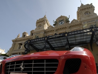 A luxury sports car with a bright red exterior is parked in front of an ornate historic building with elaborate architecture. The building has decorative statues and a large clock above its entrance, showcasing a classic architectural style.