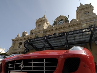 A luxury sports car with a bright red exterior is parked in front of an ornate historic building with elaborate architecture. The building has decorative statues and a large clock above its entrance, showcasing a classic architectural style.