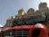 A luxury sports car with a bright red exterior is parked in front of an ornate historic building with elaborate architecture. The building has decorative statues and a large clock above its entrance, showcasing a classic architectural style.
