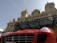 A luxury sports car with a bright red exterior is parked in front of an ornate historic building with elaborate architecture. The building has decorative statues and a large clock above its entrance, showcasing a classic architectural style.