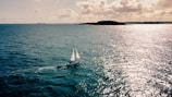 A sailboat with white sails glides across a vast, shimmering expanse of ocean under a partly cloudy sky. The sun casts bright reflections on the water, and an island is visible on the horizon.