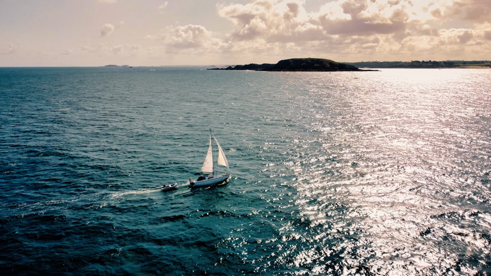 A sailboat with white sails glides across a vast, shimmering expanse of ocean under a partly cloudy sky. The sun casts bright reflections on the water, and an island is visible on the horizon.