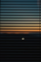 Horizontal window blinds partially cover the view of a sunset or sunrise, showcasing a gradient of colors from dark blue to light orange. A circular object, possibly the moon or sun, is visible in the lower half of the image.