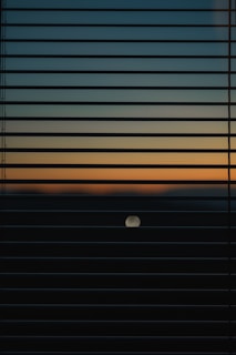Horizontal window blinds partially cover the view of a sunset or sunrise, showcasing a gradient of colors from dark blue to light orange. A circular object, possibly the moon or sun, is visible in the lower half of the image.