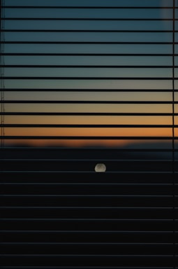 Horizontal window blinds partially cover the view of a sunset or sunrise, showcasing a gradient of colors from dark blue to light orange. A circular object, possibly the moon or sun, is visible in the lower half of the image.