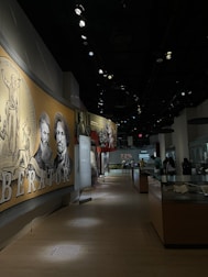 A dimly lit museum exhibit features a large mural with historical figures and text in the foreground. Several display cases line the hallway where visitors are observing various exhibits. The setting conveys an educational and historical atmosphere.