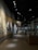 A dimly lit museum exhibit features a large mural with historical figures and text in the foreground. Several display cases line the hallway where visitors are observing various exhibits. The setting conveys an educational and historical atmosphere.
