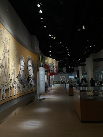 A dimly lit museum exhibit features a large mural with historical figures and text in the foreground. Several display cases line the hallway where visitors are observing various exhibits. The setting conveys an educational and historical atmosphere.
