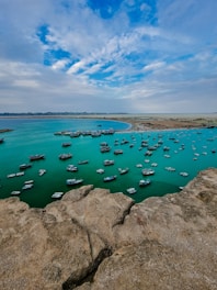 The image depicts a coastal bay area with a large number of small boats anchored in the turquoise blue water. The foreground features rugged rocks, while the background shows a sandy coastline and a partly cloudy sky.