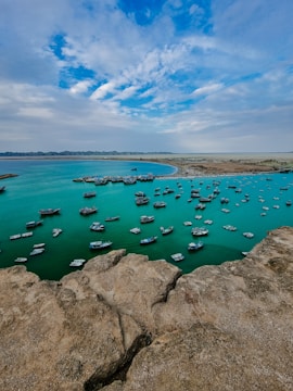 The image depicts a coastal bay area with a large number of small boats anchored in the turquoise blue water. The foreground features rugged rocks, while the background shows a sandy coastline and a partly cloudy sky.