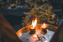 Two small, lit clay lamps placed on a metallic tray with a symbol, likely on a festive or religious occasion. The background appears to have decorative items possibly made of flowers and beads.