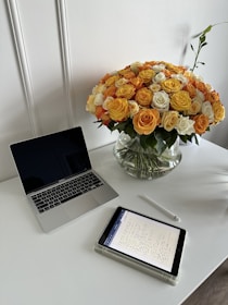A clean and modern workspace featuring a closed laptop, an electronic tablet displaying handwritten notes, and a stylus pen on a white table. A large, vibrant bouquet of yellow, orange, and white roses in a glass vase adds a colorful and fresh element to the setting.