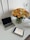 A clean and modern workspace featuring a closed laptop, an electronic tablet displaying handwritten notes, and a stylus pen on a white table. A large, vibrant bouquet of yellow, orange, and white roses in a glass vase adds a colorful and fresh element to the setting.