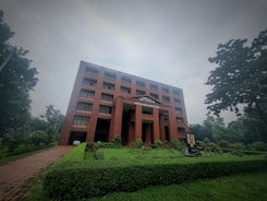 A large red brick building with multiple windows stands amidst lush greenery on a cloudy day. The building has a sign 'World Peace Science Research Centre' on the facade. In front of the building, there is a well-manicured lawn and a portrait displayed on a stand.