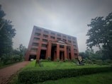 A large red brick building with multiple windows stands amidst lush greenery on a cloudy day. The building has a sign 'World Peace Science Research Centre' on the facade. In front of the building, there is a well-manicured lawn and a portrait displayed on a stand.
