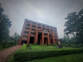 A large red brick building with multiple windows stands amidst lush greenery on a cloudy day. The building has a sign 'World Peace Science Research Centre' on the facade. In front of the building, there is a well-manicured lawn and a portrait displayed on a stand.