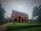 A large red brick building with multiple windows stands amidst lush greenery on a cloudy day. The building has a sign 'World Peace Science Research Centre' on the facade. In front of the building, there is a well-manicured lawn and a portrait displayed on a stand.