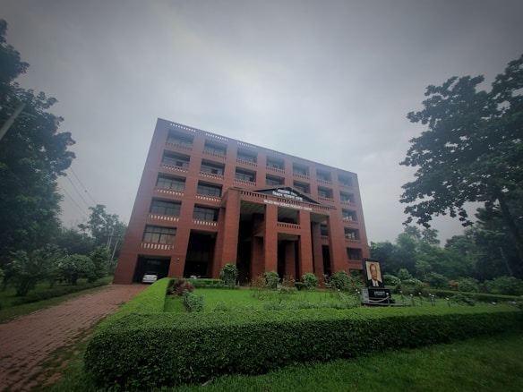 A large red brick building with multiple windows stands amidst lush greenery on a cloudy day. The building has a sign 'World Peace Science Research Centre' on the facade. In front of the building, there is a well-manicured lawn and a portrait displayed on a stand.