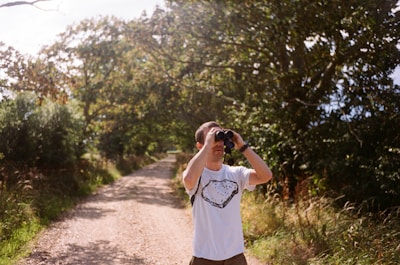 A person stands on a dirt path surrounded by lush greenery, holding binoculars up to their eyes. The path is lined with trees and bushes, and sunlight filters through the leaves, creating a serene and natural atmosphere.