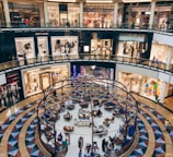 A bustling shopping mall interior with multiple high-end fashion stores like Saint Laurent and Valentino on the upper levels. The lower floor features a spherical cafe with patrons seated at tables, creating a lively atmosphere. The architecture is modern with sleek lines and glass railings.