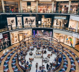 A bustling shopping mall interior with multiple high-end fashion stores like Saint Laurent and Valentino on the upper levels. The lower floor features a spherical cafe with patrons seated at tables, creating a lively atmosphere. The architecture is modern with sleek lines and glass railings.