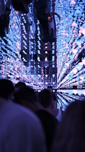 A futuristic tunnel is adorned with luminous lights resembling jellyfish, extending into the distance. The reflective surfaces create an illusion of endless space, with people walking through, partially silhouetted against the vibrant backdrop. The atmosphere mixes mystery with a sense of exploration.