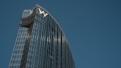 A modern skyscraper with a sleek glass facade reflecting the sky. The building's design features a curved structure with the letter 'W' prominently displayed at the top. The blue sky serves as a backdrop, enhancing the architectural elegance.