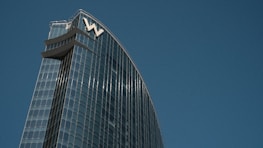 A modern skyscraper with a sleek glass facade reflecting the sky. The building's design features a curved structure with the letter 'W' prominently displayed at the top. The blue sky serves as a backdrop, enhancing the architectural elegance.