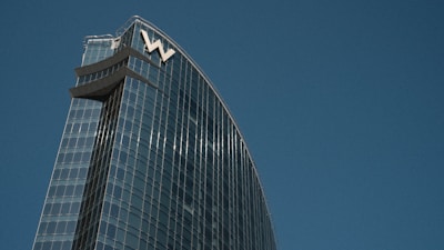 A modern skyscraper with a sleek glass facade reflecting the sky. The building's design features a curved structure with the letter 'W' prominently displayed at the top. The blue sky serves as a backdrop, enhancing the architectural elegance.