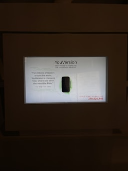 A digital display features the YouVersion Bible app, highlighting its significance in changing how people read the Bible. The display includes an image of the first iPhone used to download the app, along with a quote from The New York Times. It also shows a statistic about the number of Bible plans completed, emphasizing the wide reach and usage of the app.