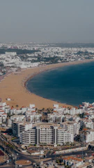A coastal cityscape featuring a sandy beach stretching along a curving shoreline. The beach is lined with a multitude of white buildings with red roofs, indicative of a residential or tourist area. The vibrant blue sea contrasts with the golden sand, and a few beachgoers are visible enjoying the sunny day.