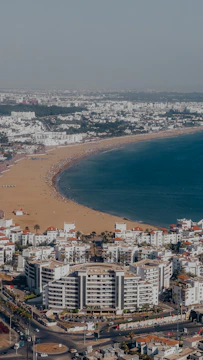 A coastal cityscape featuring a sandy beach stretching along a curving shoreline. The beach is lined with a multitude of white buildings with red roofs, indicative of a residential or tourist area. The vibrant blue sea contrasts with the golden sand, and a few beachgoers are visible enjoying the sunny day.
