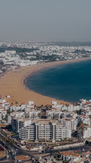 A coastal cityscape featuring a sandy beach stretching along a curving shoreline. The beach is lined with a multitude of white buildings with red roofs, indicative of a residential or tourist area. The vibrant blue sea contrasts with the golden sand, and a few beachgoers are visible enjoying the sunny day.