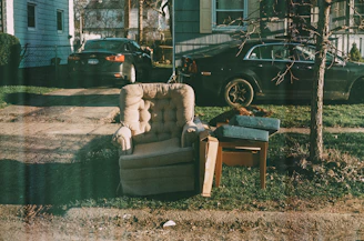An upholstered armchair sits on a patch of grass next to a small wooden table with some cushions and a tree nearby. Two parked cars are visible in the driveway of a house, with a leafless tree casting shadows on the ground.