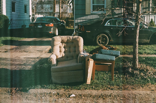 An upholstered armchair sits on a patch of grass next to a small wooden table with some cushions and a tree nearby. Two parked cars are visible in the driveway of a house, with a leafless tree casting shadows on the ground.